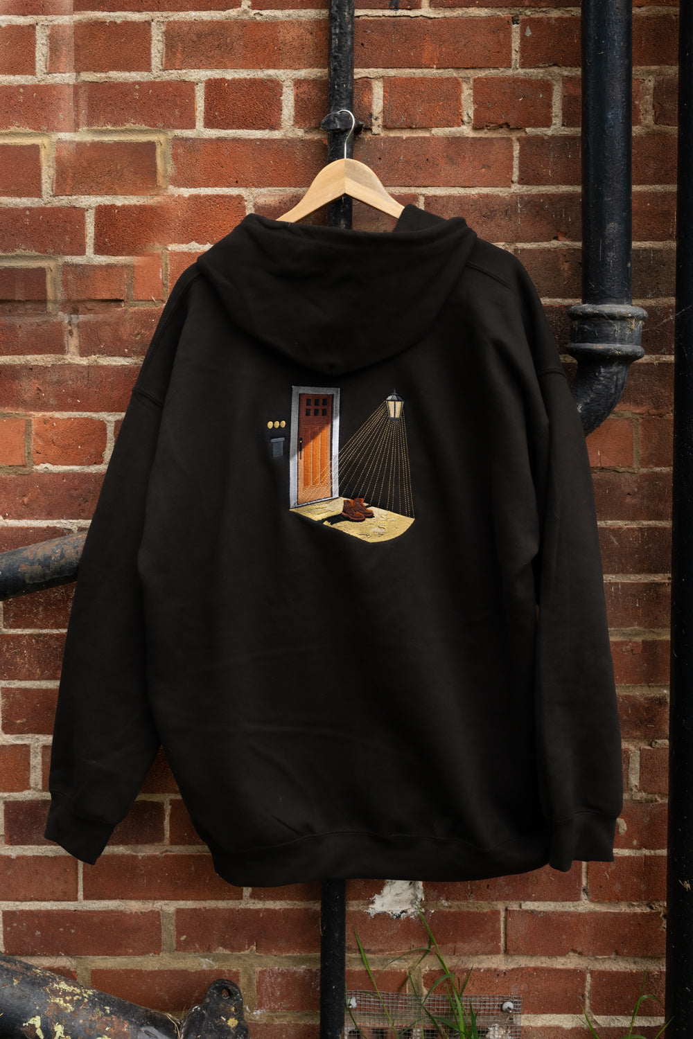 Back view of black zip-up hoodie on a hanger, large embroidered porch scene with open door, glowing lamp, and boots on doorstep - Noah Kahan Porch Light inspired, The Great Divide merch
