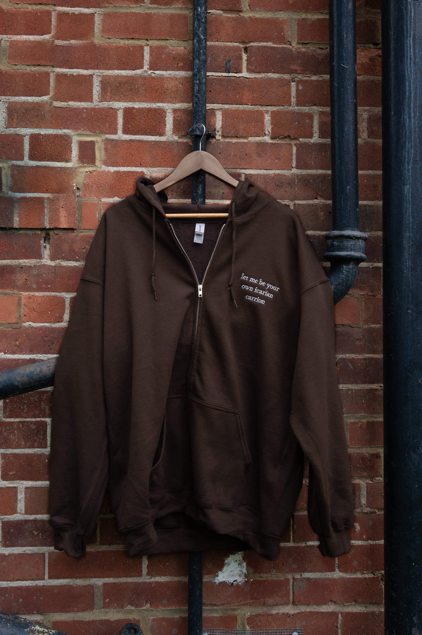 Brown fleece-lined zip-up hoodie on a hanger showing silver metal zipper hardware and white thread lyric embroidery "let me be your own icarian carrion" from Unreal Unearth by Hozier.