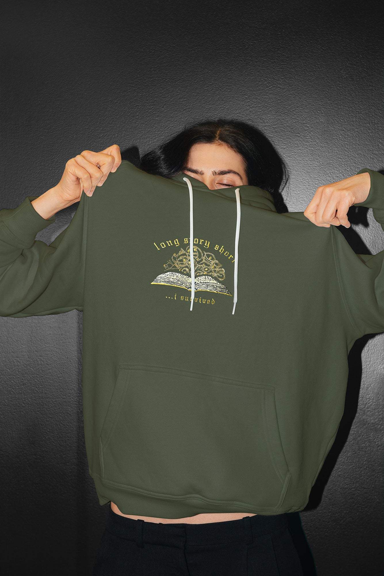 Person wearing the embroidered "long story short, i survived" hoodie with book illustration in olive. Inspired by Taylor Swift's evermore.