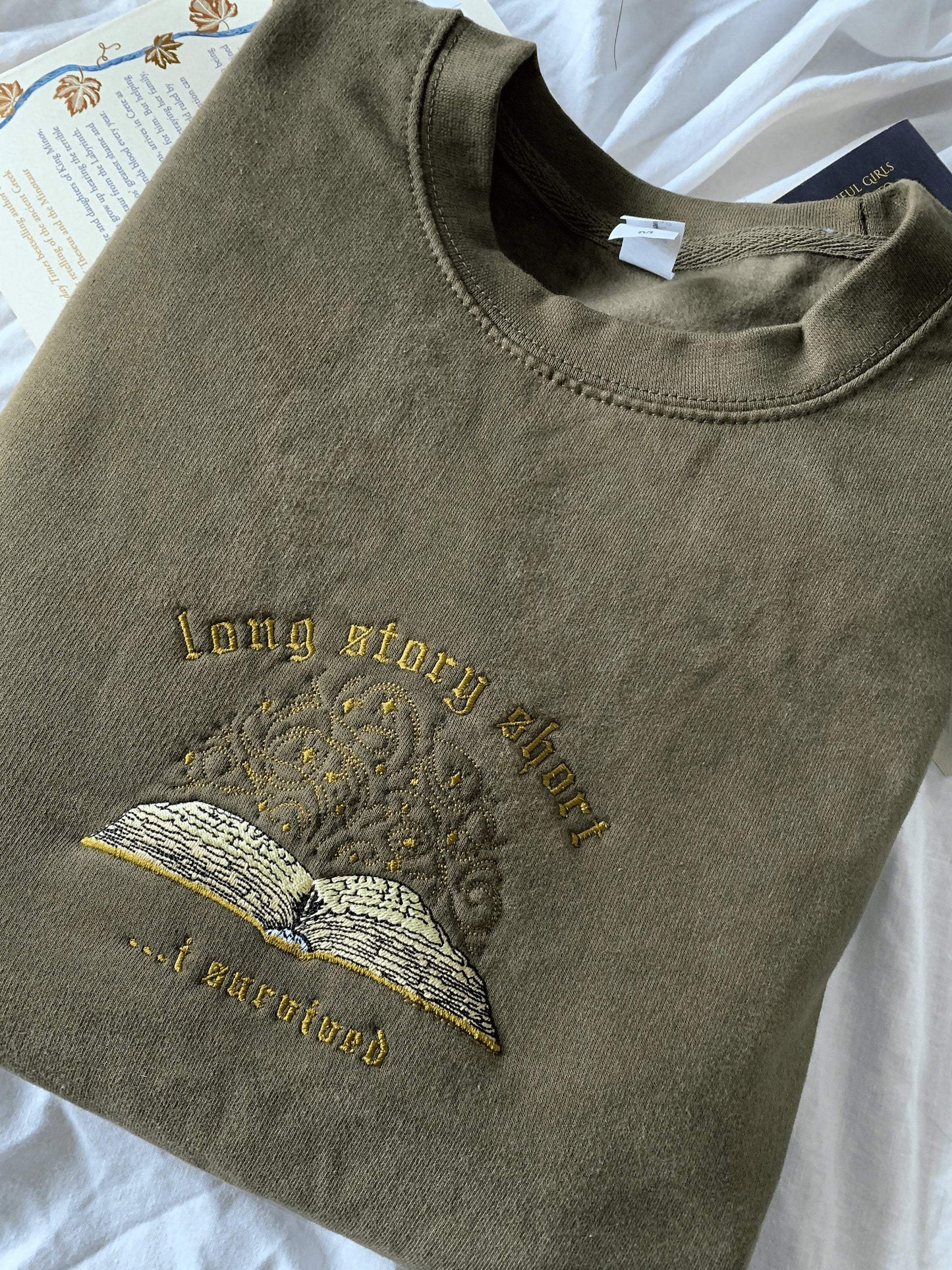Hand-drawn embroidered "long story short, i survived" sweatshirt with book illustration, inspired by Taylor Swift's evermore. Olive flat lay.
