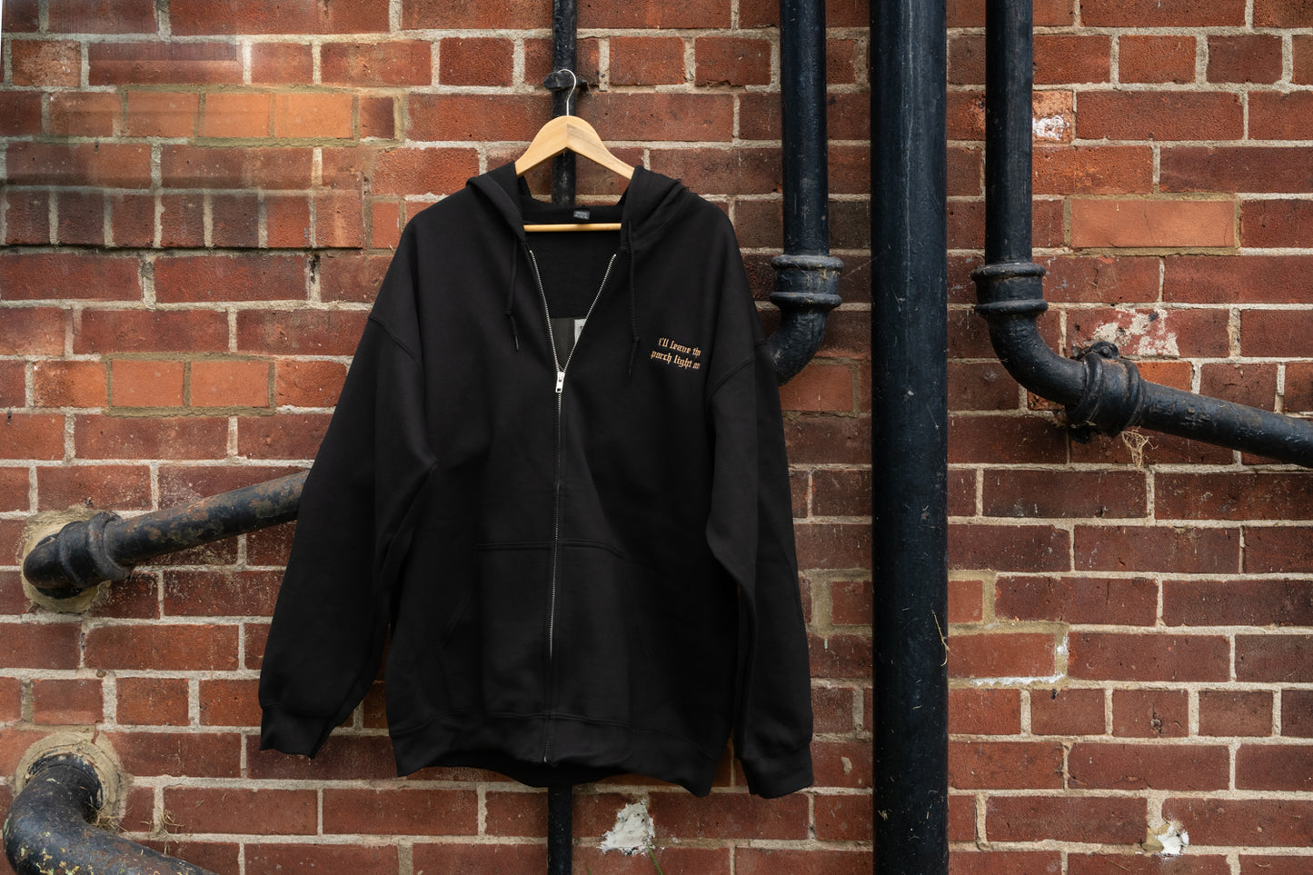 Black zip-up hoodie on a hanger, full-zip silver metal closure, drawstring hood, kangaroo pouch pockets, small left-chest lyric embroidery - unisex fit, S–5XL sizing