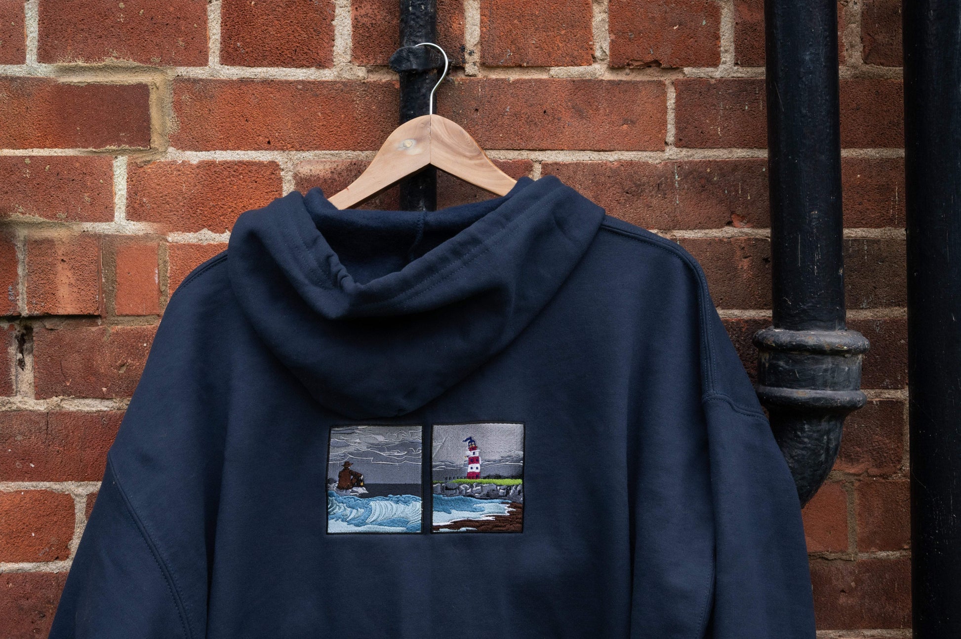 Back view of navy fleece zip-up hoodie on a hanger, two embroidered panels showing a figure with lantern on stormy rocks beside a red and white lighthouse on a coastal outcrop - Lord Huron Ghost on the Shore inspired embroidered hoodie

