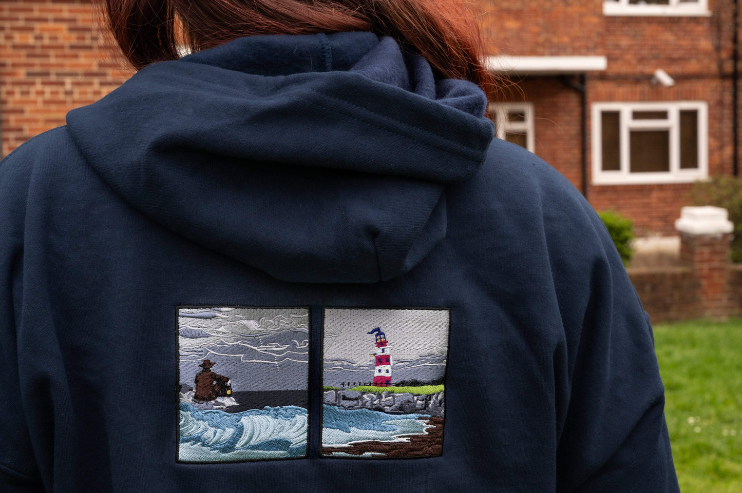 Close-up of dual embroidered panels on navy hoodie back - left panel: lone figure with lantern on rocks in crashing waves under stormy sky; right panel: red and white striped lighthouse on coastal rocks. Inspired by The Ghost on The Shore by Lord Huron.