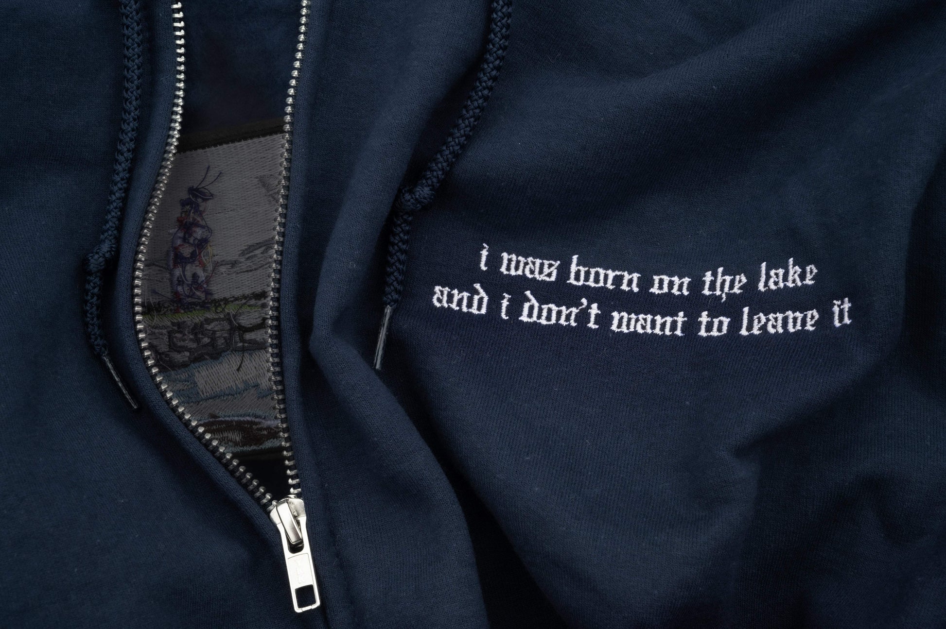 Close up of a full-zip fleece hoodie, silver metal zipper, drawstring hood, small left-chest old-English lyric embroidery reading "i was born on the lake and i don't want to leave it".