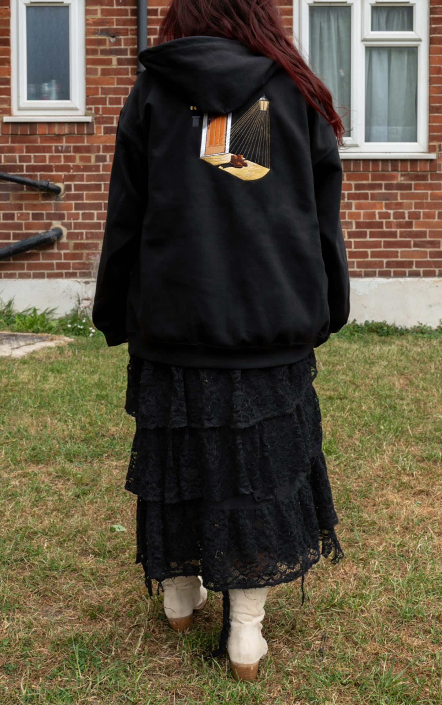 Back view of black zip-up hoodie worn over a tiered black skirt with white cowboy boots outdoors, large Porch Light embroidery visible. Styled as a warm fleece-lined layer for a cold Noah Kahan outdoor concert.