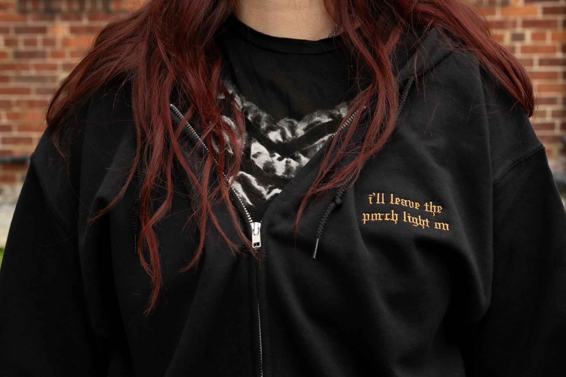 Worn close-up of a model wearing the Porch Light fleece lined black zip-up hoodie featuring a silver metal zipper, drawstring hood, small left-chest old-English lyric embroidery.

