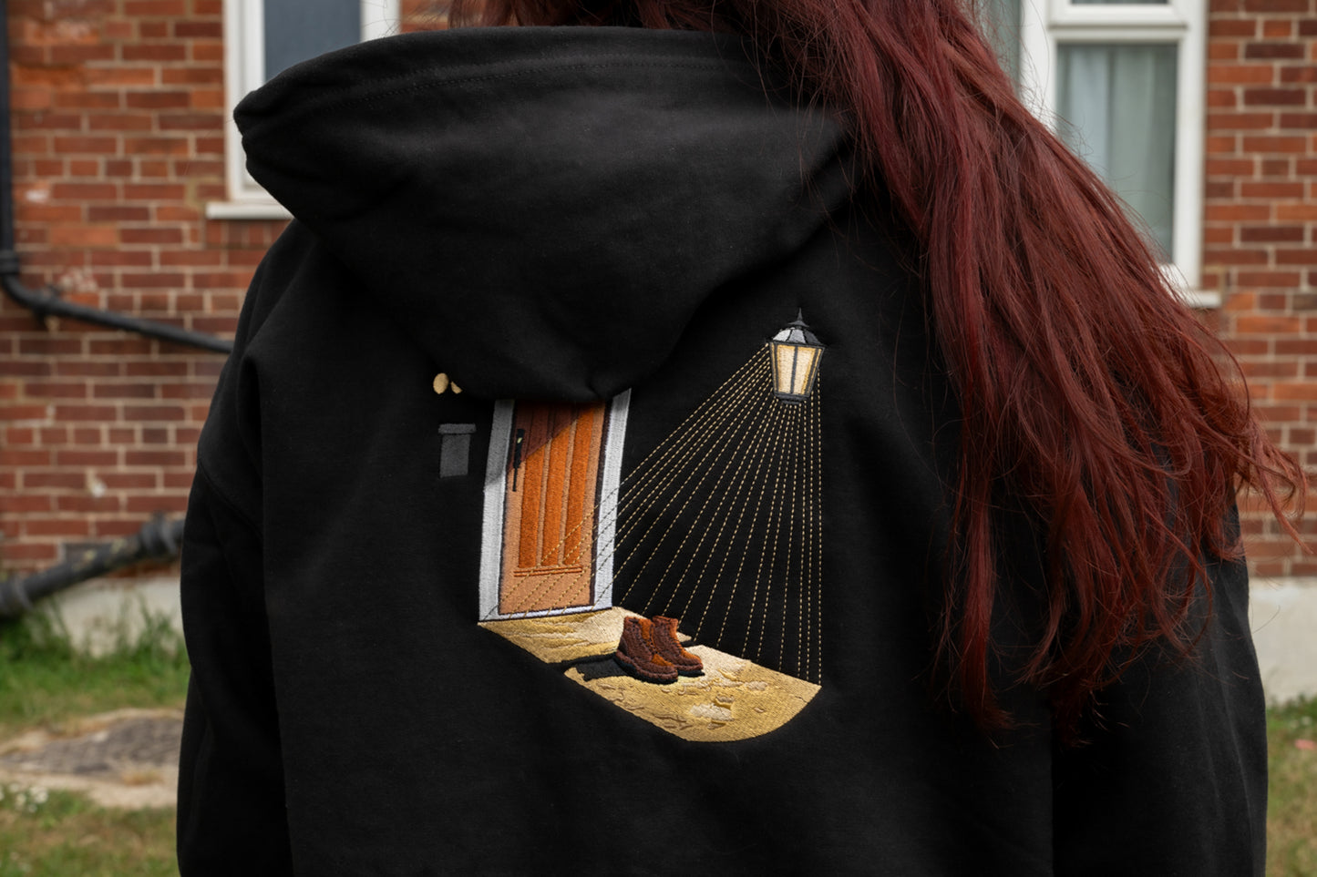 Worn close-up of Porch Light back embroidery on black zip-up hoodie - detailed scene of open door, lamp light, and boots on a threshold in amber, brown, and gold tones on black fleece-lined fabric.