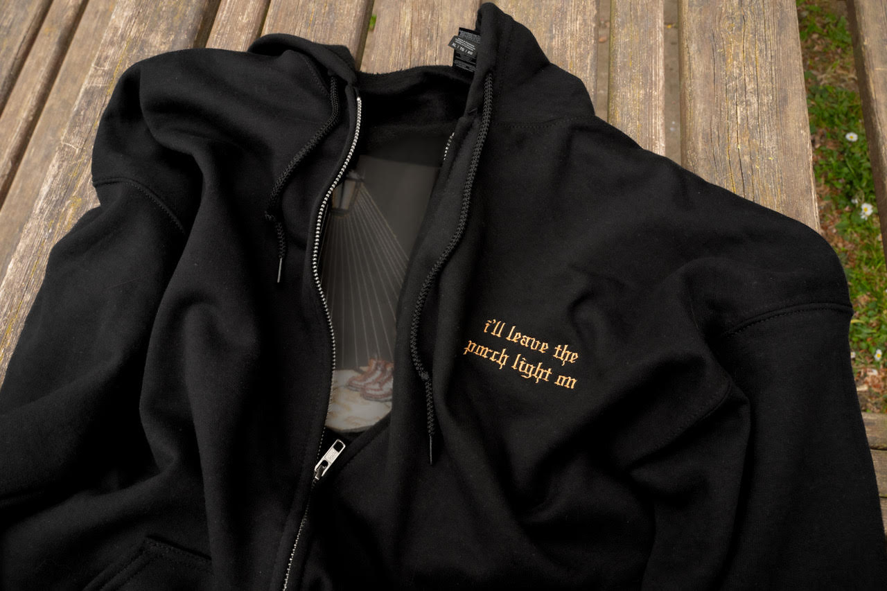 Flat lay front view of the black Porch Light embroidered zip-up hoodie with drawstring hood, silver zipper, pouch pockets, and small lyric embroidery on the left chest.