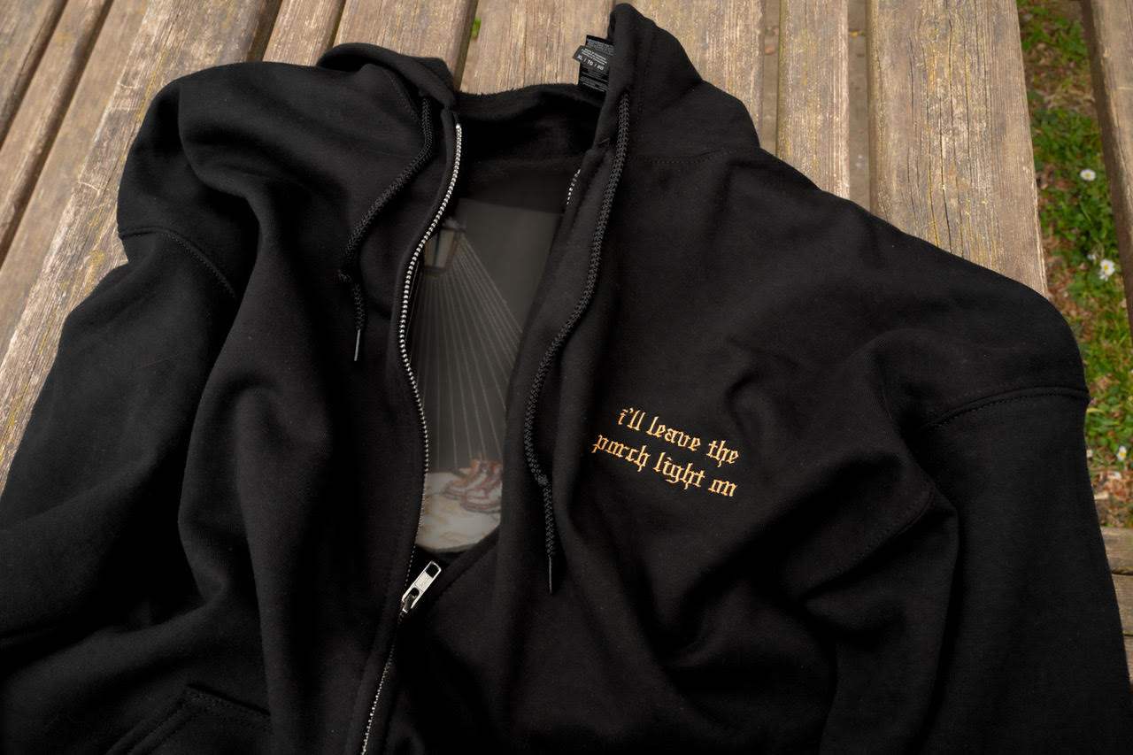 Flat lay front view of the black Porch Light embroidered zip-up hoodie with drawstring hood, silver zipper, pouch pockets, and small lyric embroidery on the left chest.