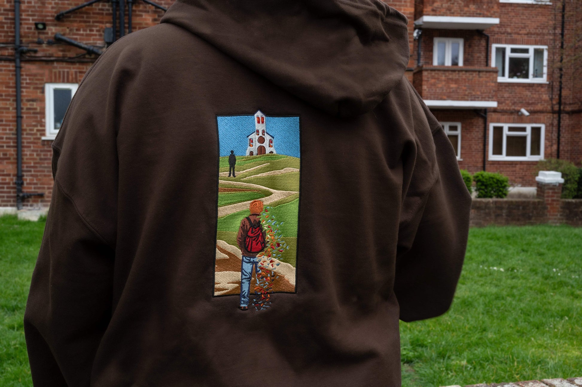 Worn close-up of The Great Divide back embroidery on brown zip-up hoodie -winding path, church on a hill, and stained glass fragments falling from a walking figure, stitched in greens, blues, and earth tones on brown fleece-lined fabric